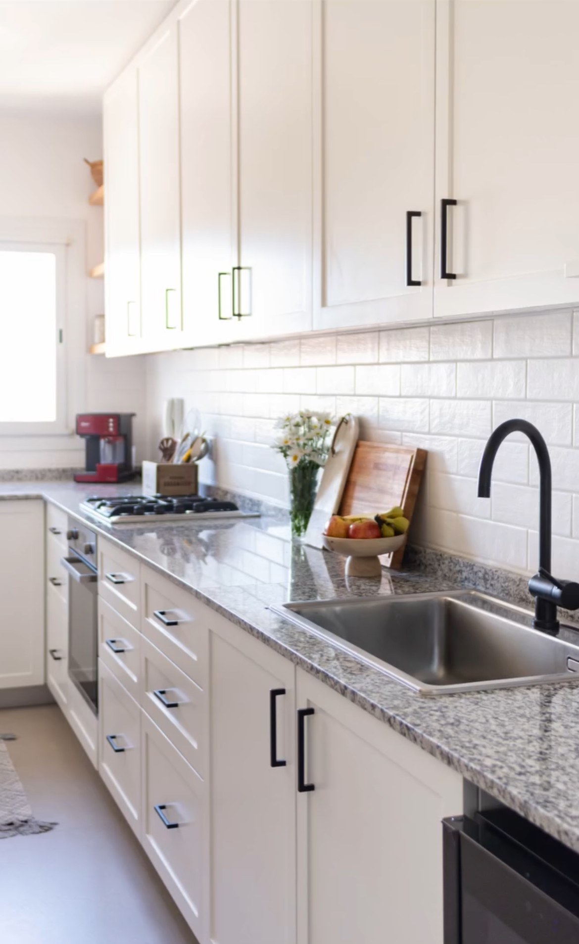 Cozinha com bancada de granito Branco Fortaleza, cinza claro salpicado, cuba de inox, fruteira e tábua de madeira junto ao azulejo branco