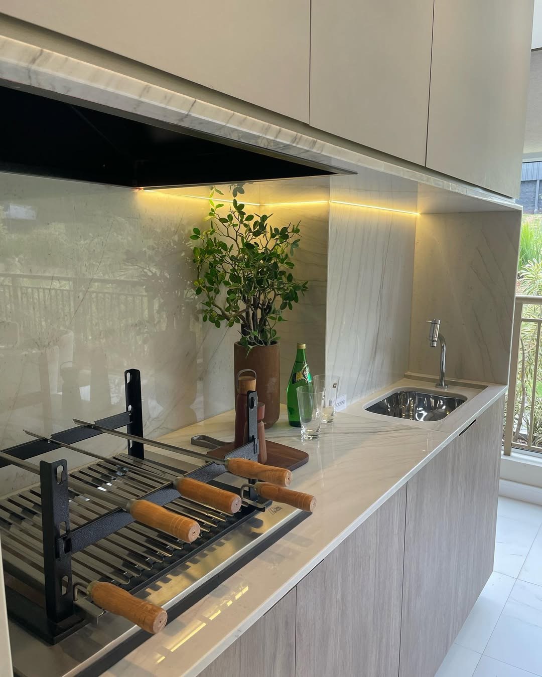 Bancada clara de quartzito Denali com cuba de inox, garrafas, copos e vaso com planta junto a uma parede com veios suaves