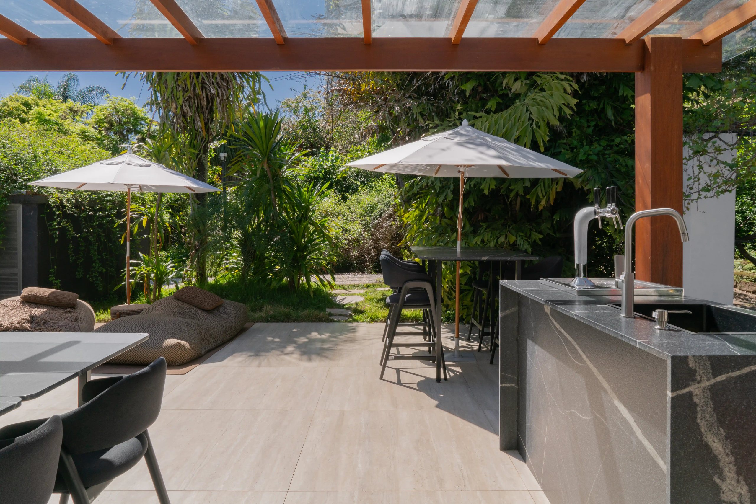 Área gourmet externa com bancada e cuba em granito Vincent Black, piso claro, mesas com ombrelones e jardim tropical ao fundo
