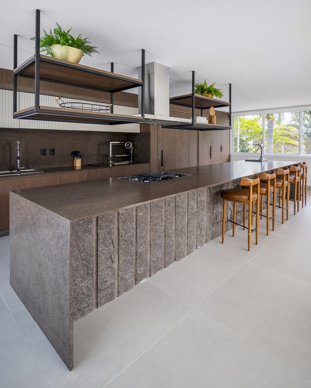 Cozinha com ilha longa em granito Café Imperial descendo em cascata na lateral, banquetas de madeira, cooktop e prateleira suspensa