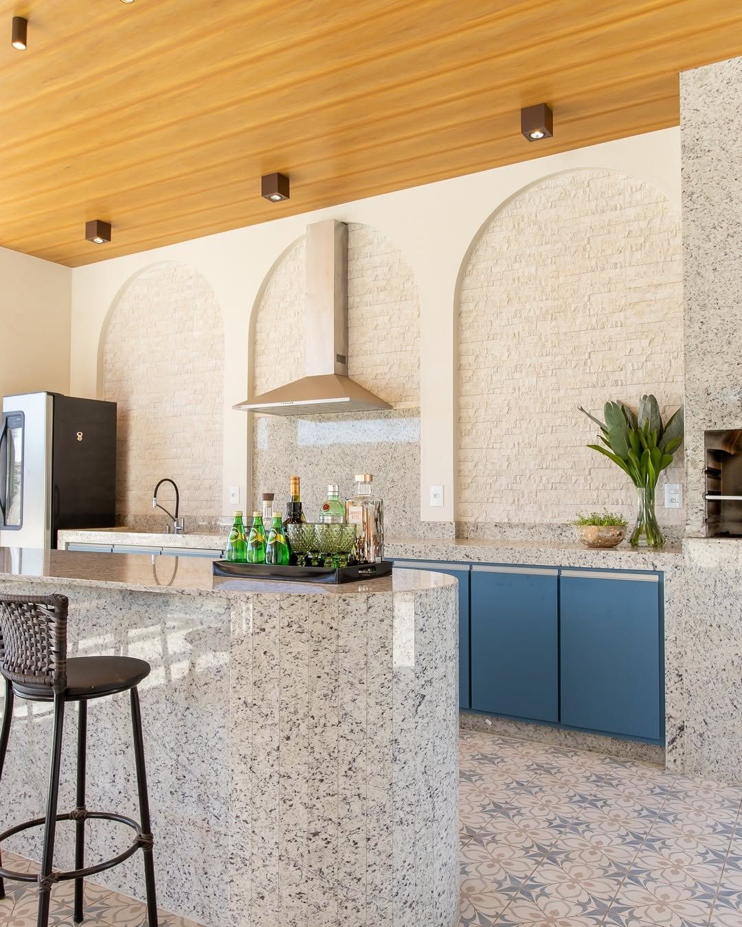Área gourmet com ilha curva e bancada em granito Dallas White, coifa, parede de tijolos claros com arcos e armários azuis