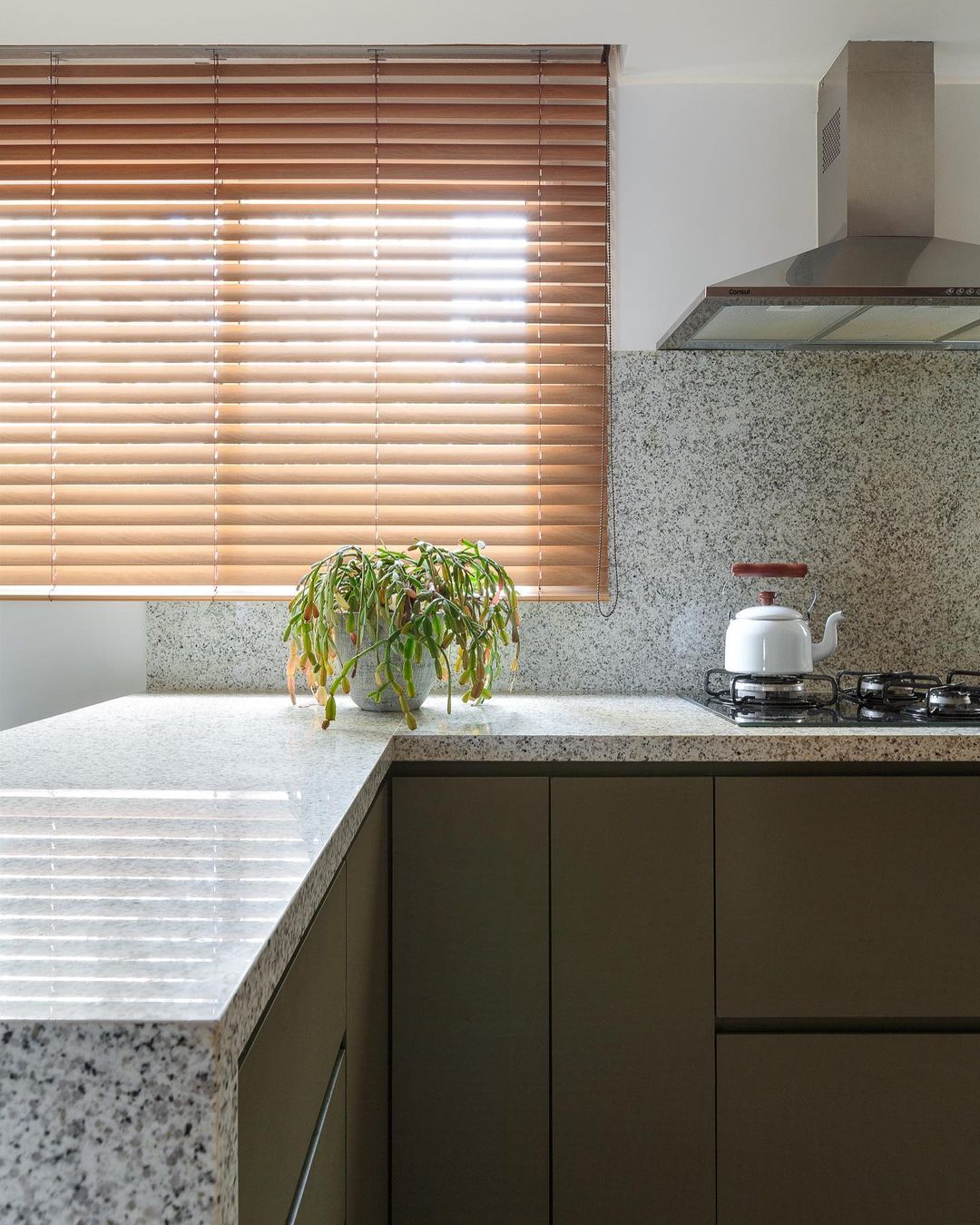 Cozinha com bancada em L e frontão em granito Branco Ceará, cooktop, chaleira branca, vaso de planta e persianas de madeira